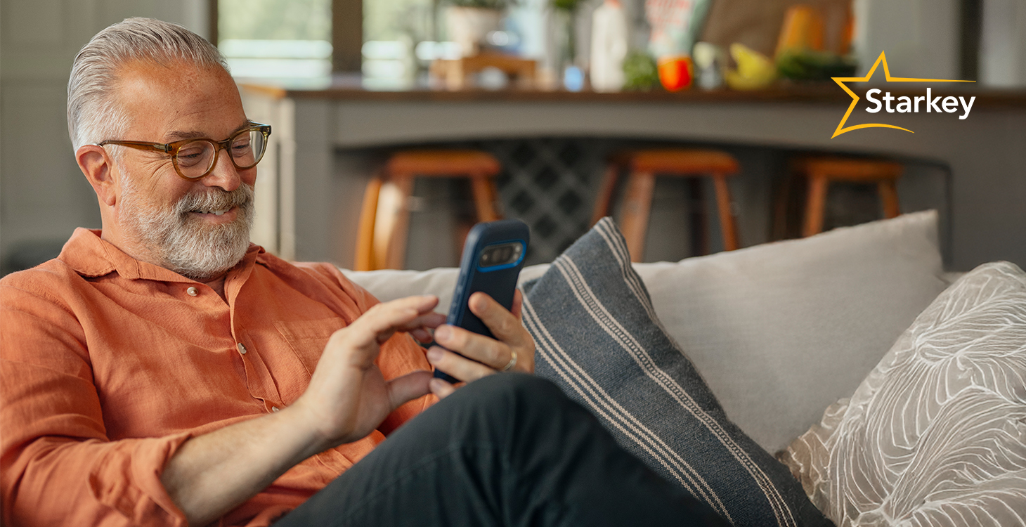 Image of smiling middle aged man sitting on his couch and typing on his smartphone
